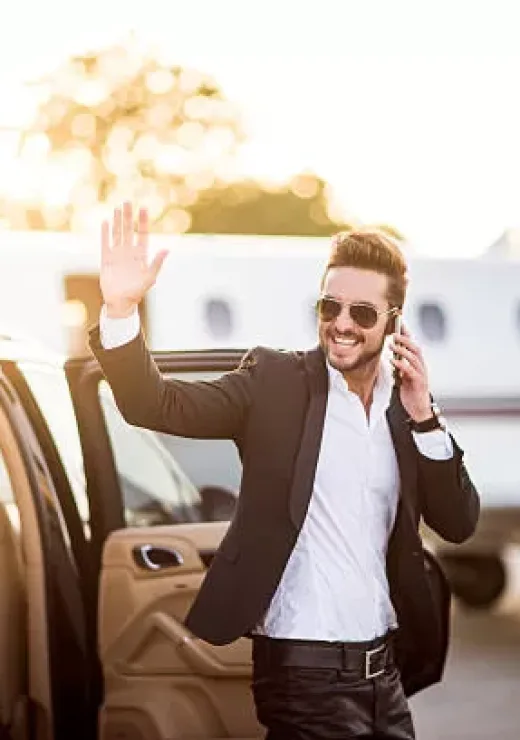 Young well dressed man with sunglasses exiting from the back seat of the car, holding his smart phone and waving to someone. He looks like a famous musician or other celebrity. Private jet airplane is in the background.